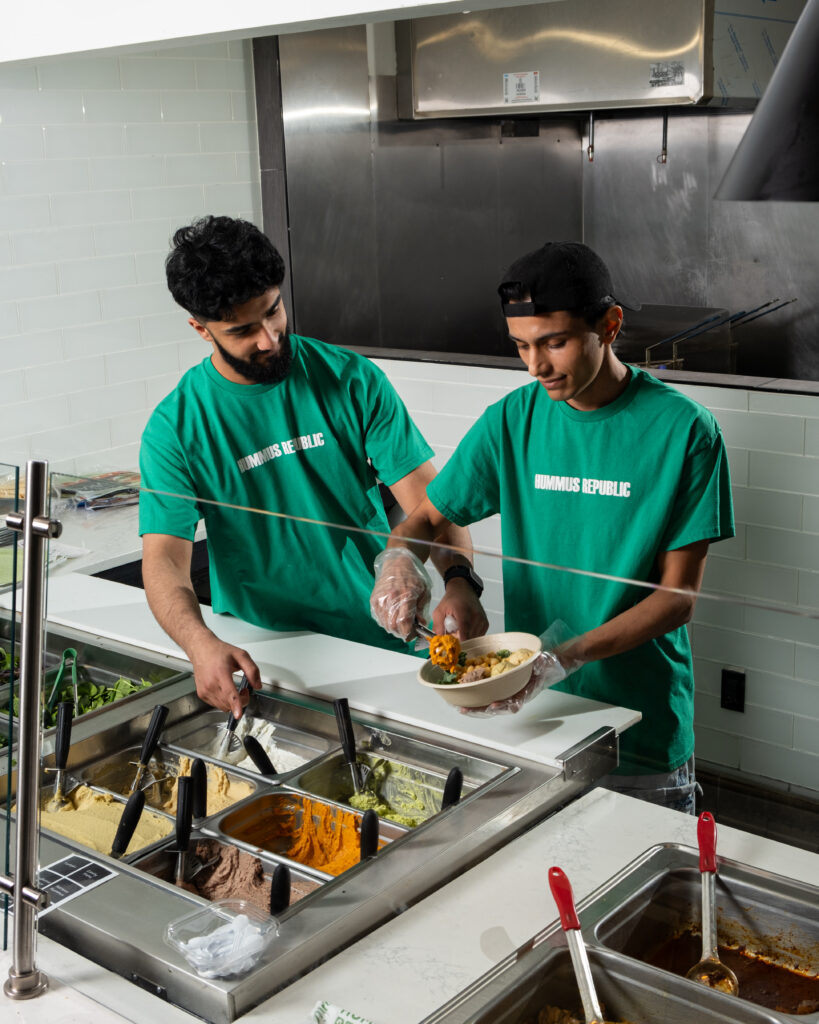 two employees in green hummus republic shirts prepare a bowl with various toppings at a food counter in a restaurant kitchen.