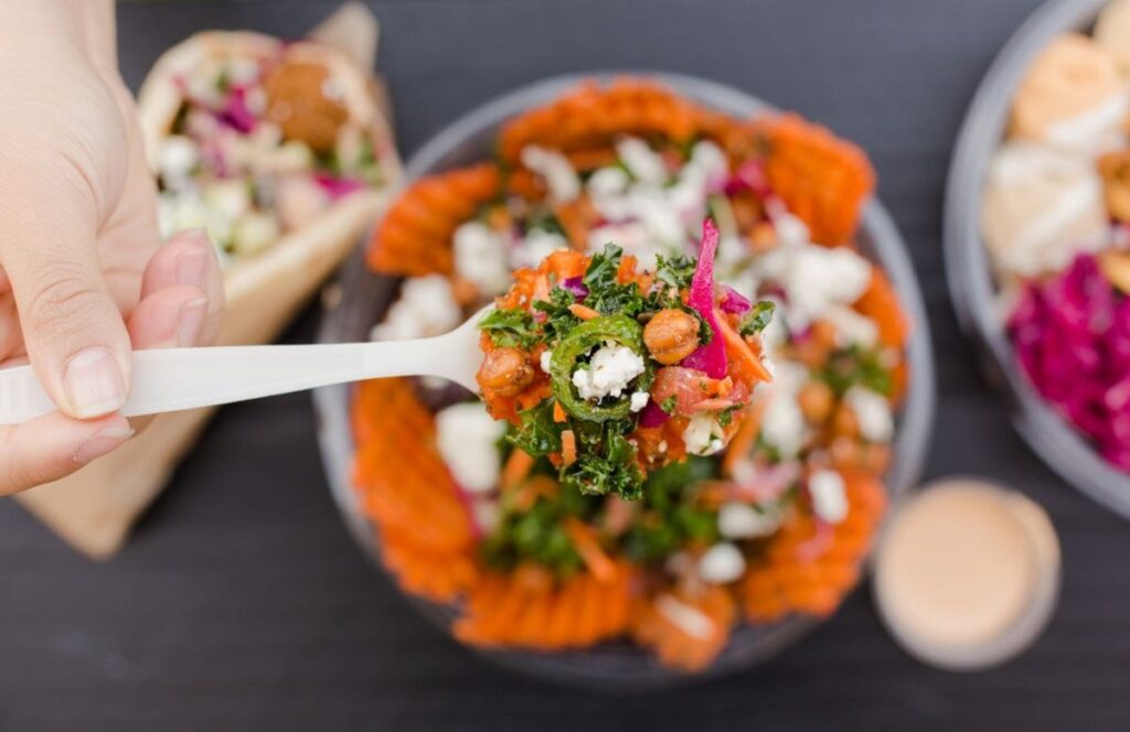 What It Actually Means to Own a Franchise Built Around Food You Already Love a hand holds a white plastic fork with salad above a bowl of mixed greens, sweet potato fries, chickpeas, feta, and vegetables. other food items are visible in the background.