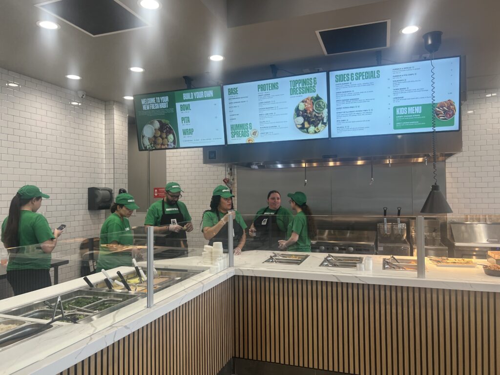 staff in green uniforms stand behind the counter of a fast casual restaurant, preparing food. digital menu boards display options above them.