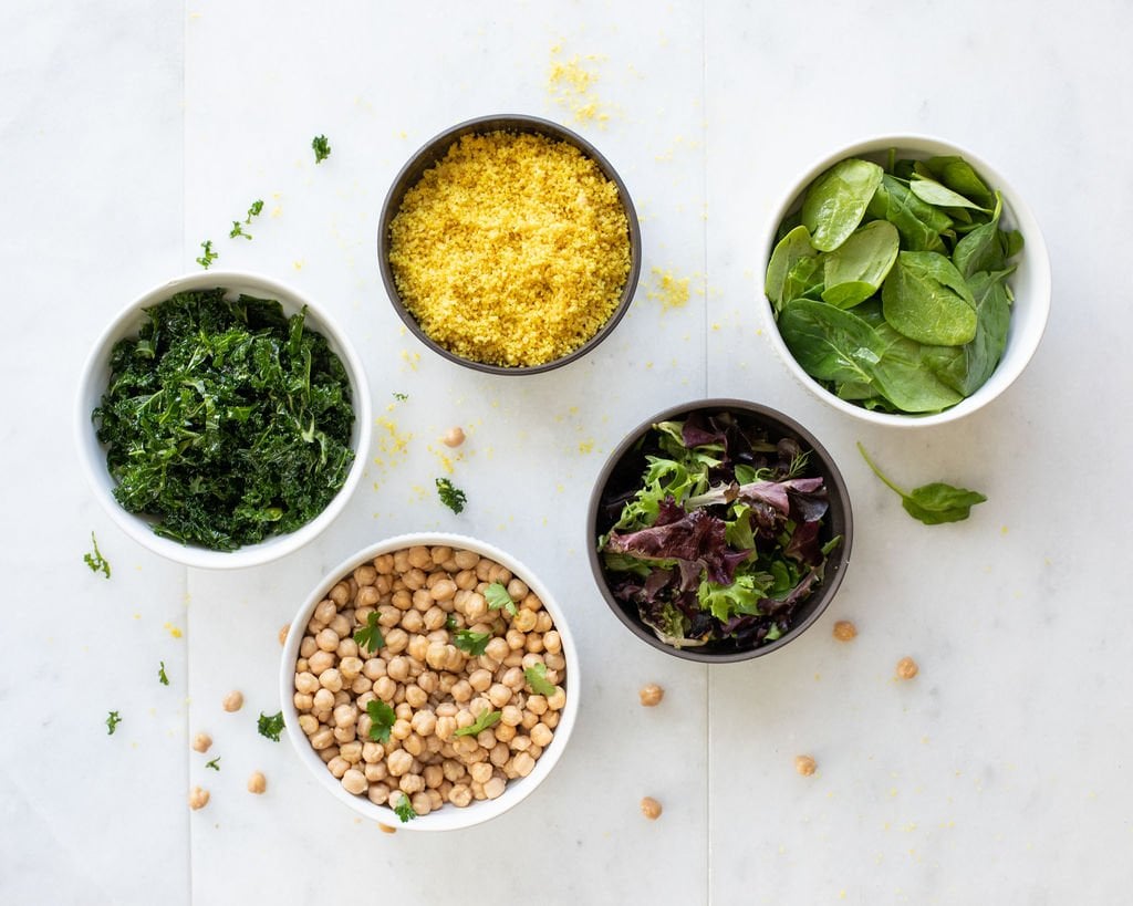 five bowls containing chopped kale, cooked couscous, fresh spinach, mixed salad greens, and chickpeas, arranged on a white surface with scattered leaves and chickpeas.