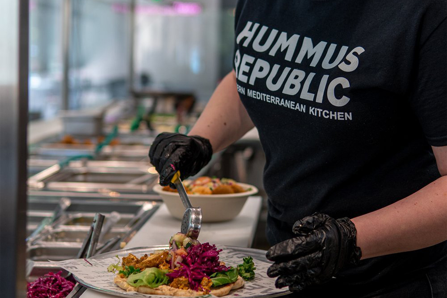 Contact a person wearing gloves and a hummus republic shirt prepares a mediterranean dish at a food counter, adding toppings to a plate.