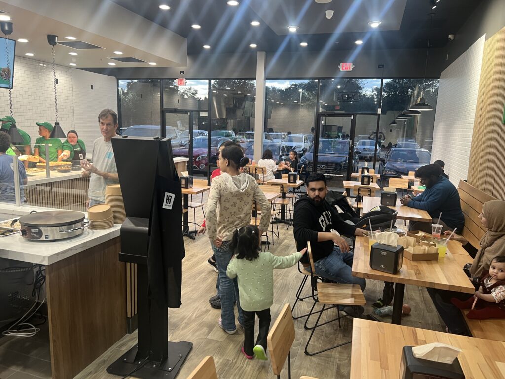 people dining and waiting in a modern, casual restaurant with large windows, wooden tables, and staff working behind the counter in the background.