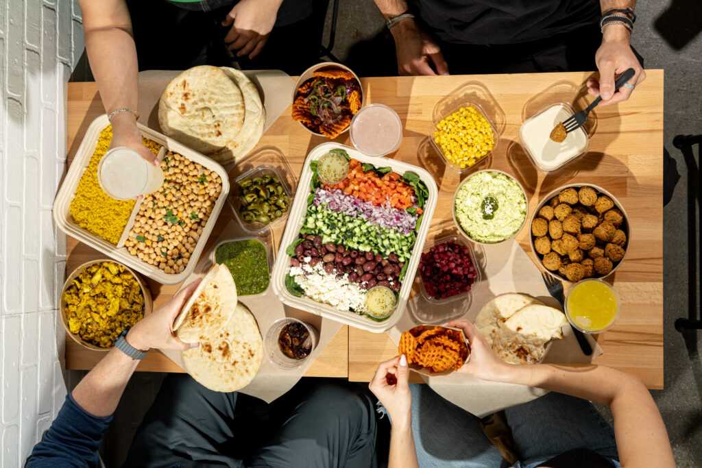 four people share a meal at a wooden table with falafel, pita, salads, grains, sauces, and drinks arranged in plastic containers.