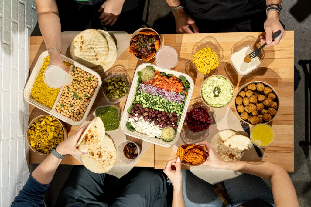 What No One Tells You About Leaving a Stable Job to Own a Business four people sharing a meal with assorted mediterranean dishes, including pita bread, falafel, salads, hummus, and various toppings, on a wooden table.