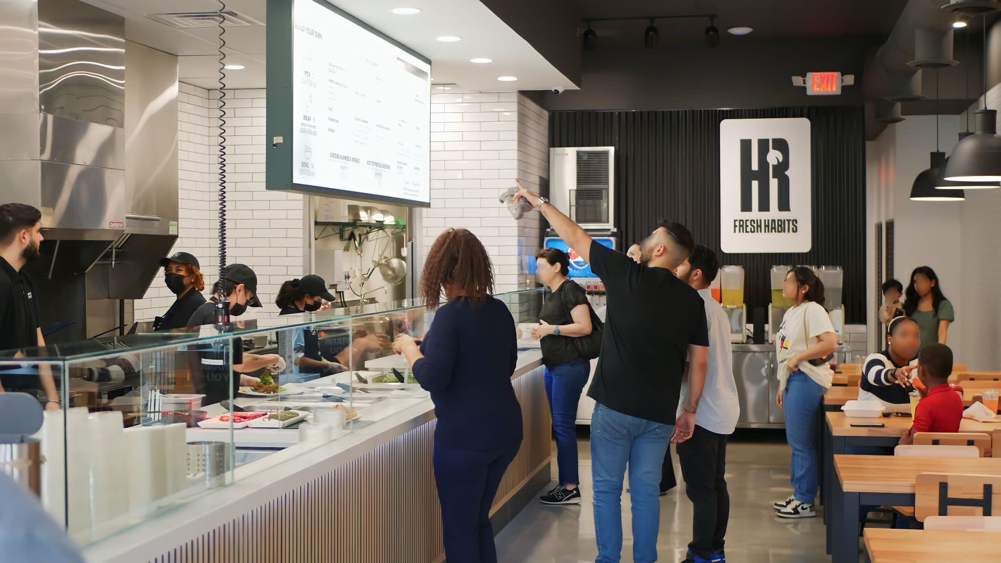 Services customers stand in line and order food at a modern counter service restaurant with an open kitchen and a menu displayed on a large screen.