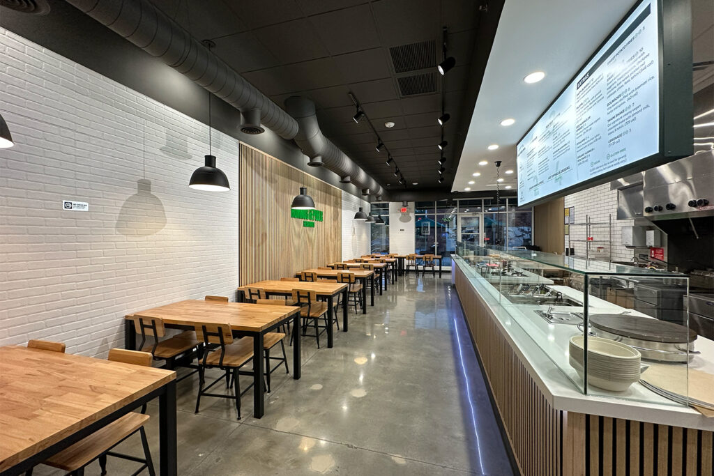 modern, well lit restaurant interior with wooden tables and chairs on the left and a food service counter with a large overhead menu on the right.