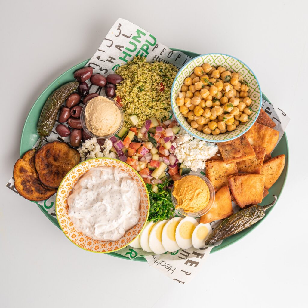 a green platter with hummus, tzatziki, pita chips, chickpeas, falafel, feta cheese, olives, sliced egg, grilled vegetables, and a chopped salad.
