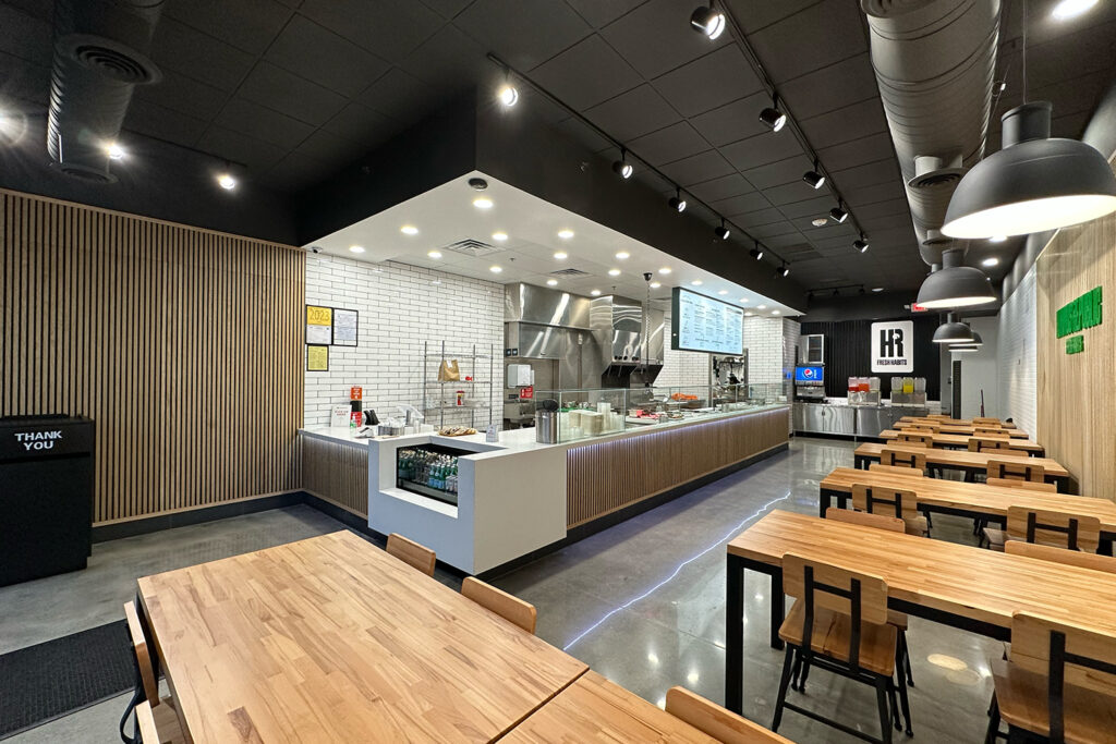 a modern, clean restaurant interior with wooden tables and chairs, a counter for food service, menu boards, and a visible kitchen area.