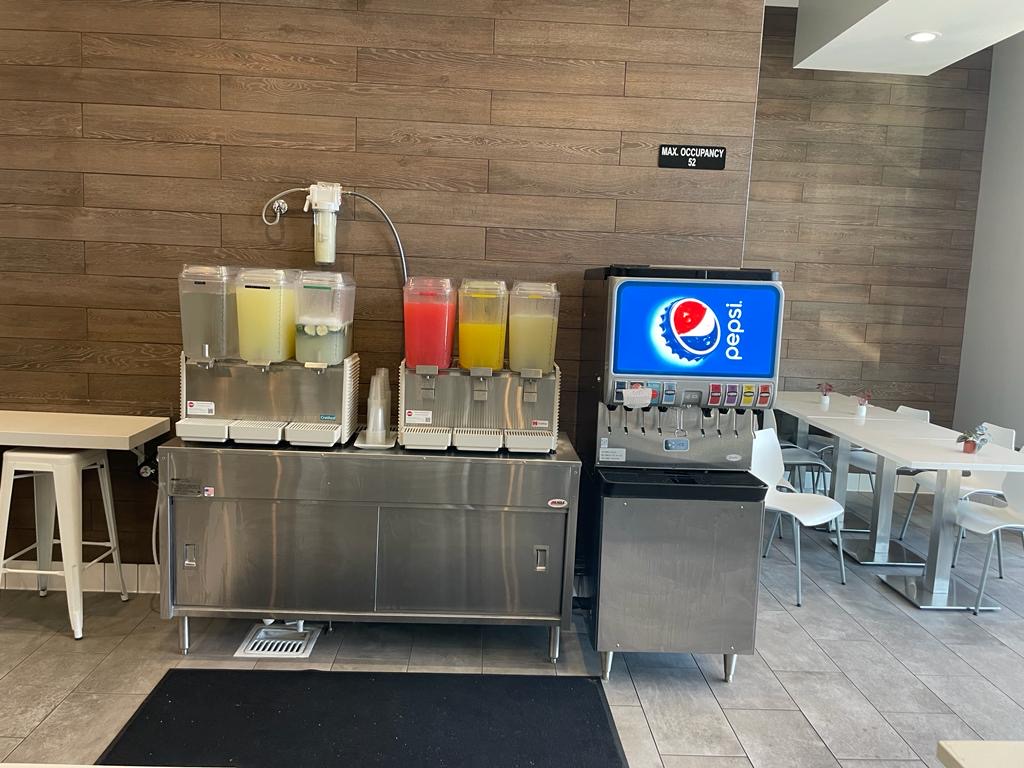 a beverage station with four juice dispensers and a pepsi soda fountain machine, set against a wood paneled wall in a dining area with white tables and chairs.