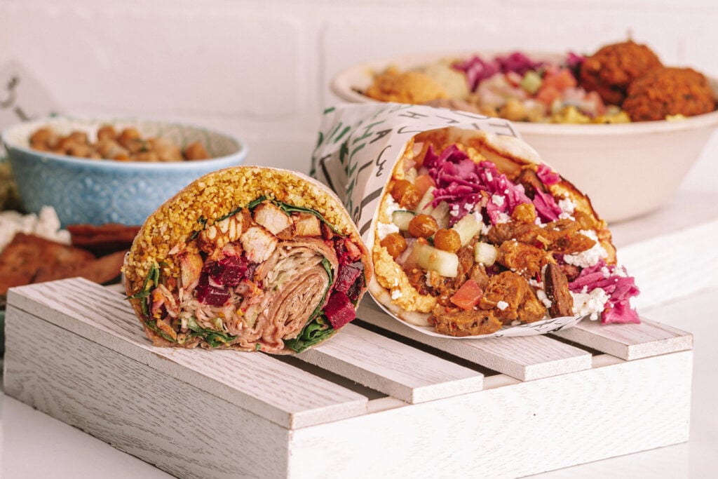 What No One Tells You About Leaving a Stable Job to Own a Business two wraps filled with vegetables, grains, and protein are placed on a wooden tray; salad bowls with toppings are visible in the background.
