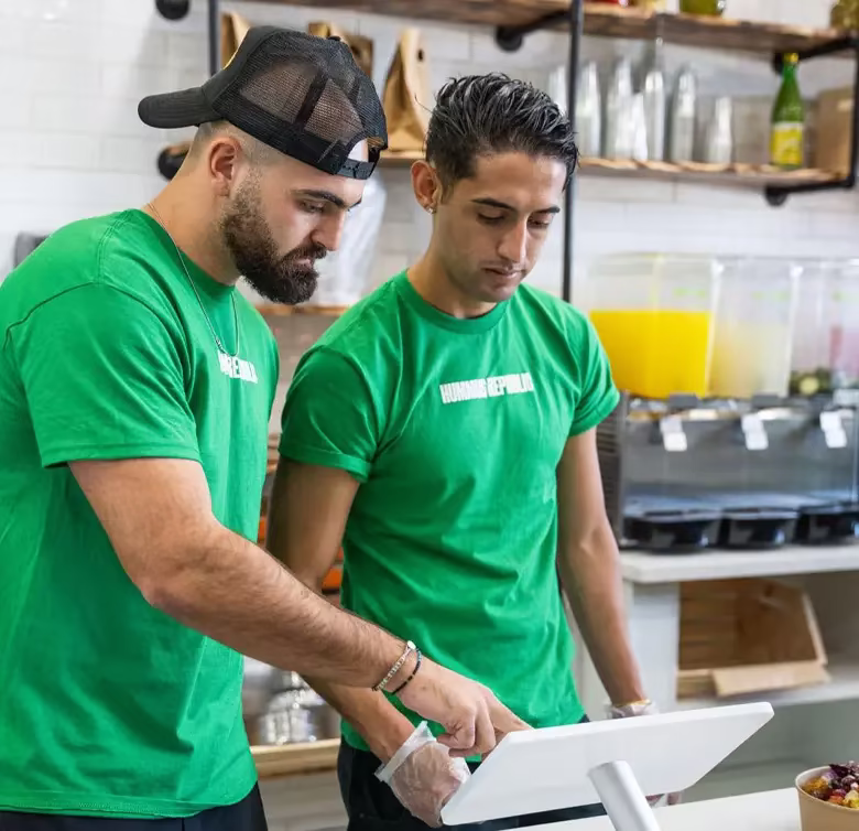 Do You Actually Need a Food Handler License to Own a Franchise Restaurant? two men in green shirts work together at a touchscreen register in a café or juice bar, with drinks and utensils visible in the background.