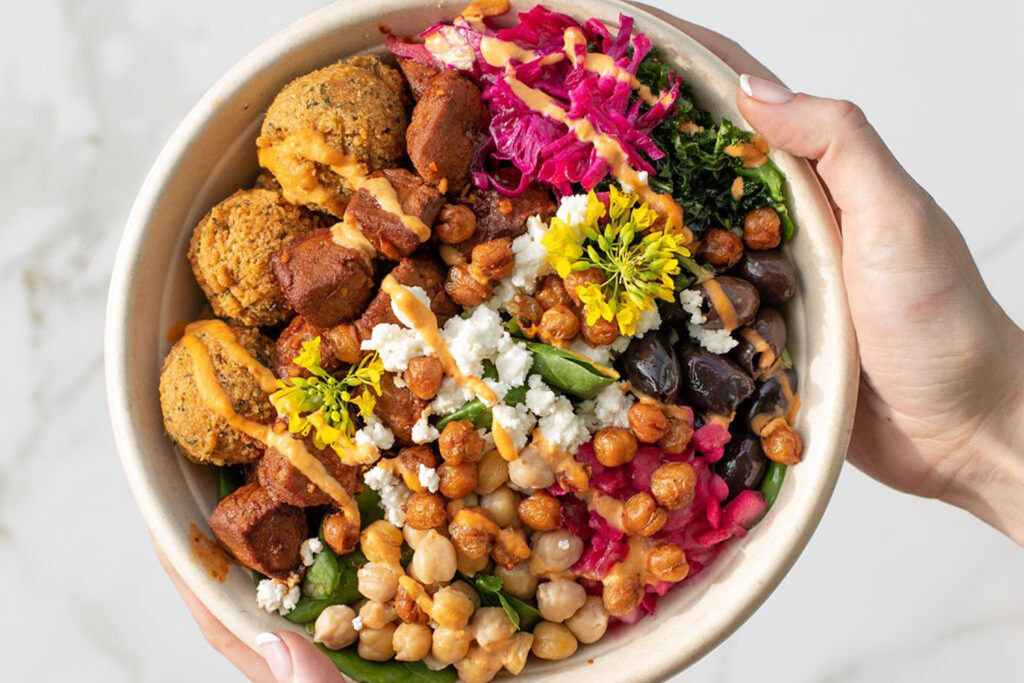 a bowl filled with falafel, chickpeas, feta cheese, olives, greens, pickled cabbage, and sauce, topped with edible flowers.