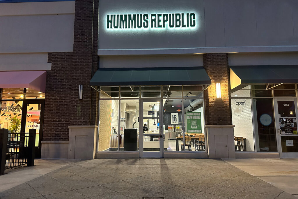 How Owning a Mediterranean Restaurant Lets You Represent Your Community — Not Just Serve It exterior of hummus republic restaurant at dusk, showing glass doors and windows, green awnings, and illuminated sign above the entrance.