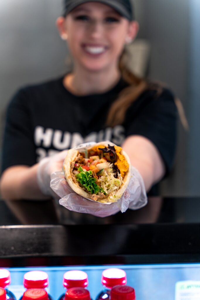 What Does It Really Cost to Open a Fast Casual Restaurant? An Honest Breakdown a person in a black shirt and cap, wearing gloves, holds out a filled pita wrap towards the camera; bottles are visible in a fridge below.
