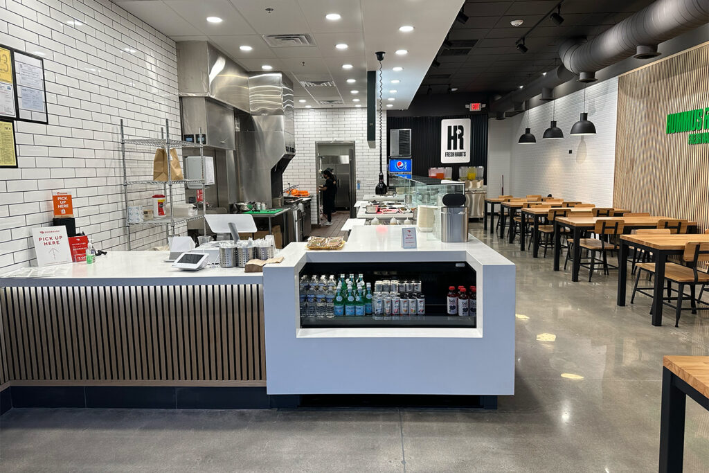 modern fast casual restaurant interior with white tiled walls, visible kitchen area, counter with drinks, and wooden tables and chairs neatly arranged for dine in customers.