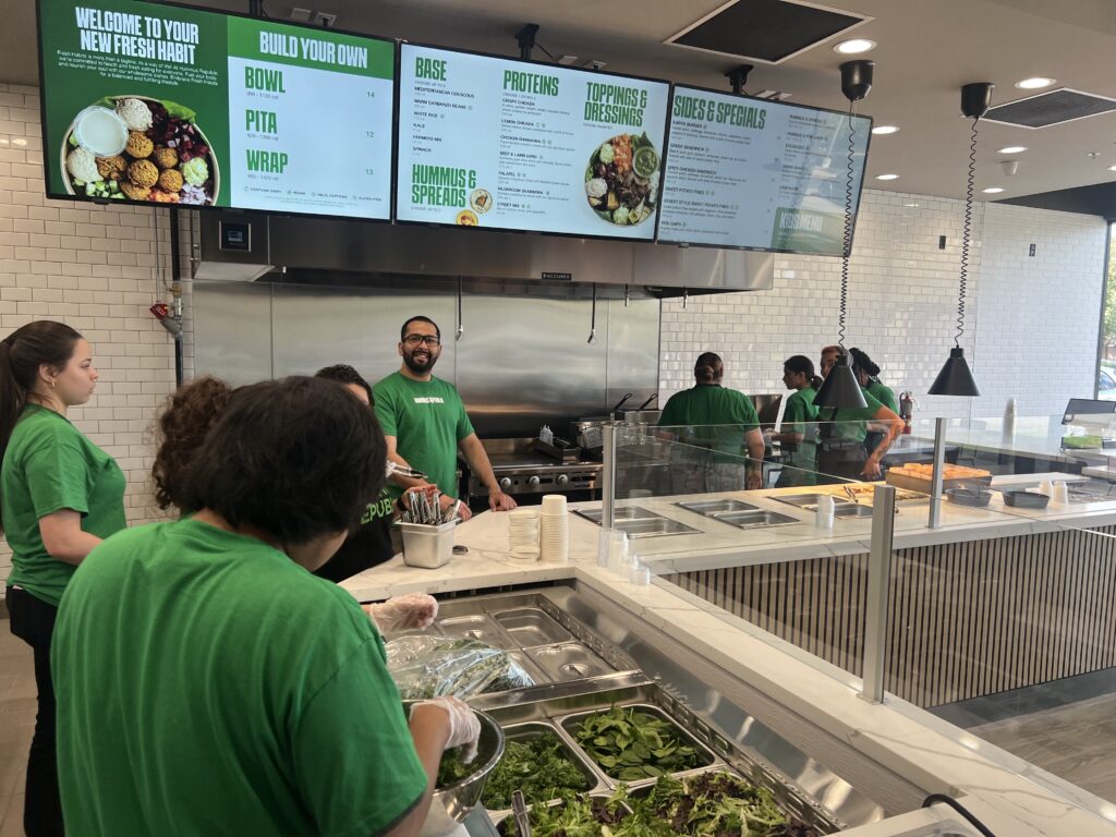 What a Manager-Run Location Model Actually Does to Your Income and Your Schedule staff in green shirts prepare food behind the counter at a fast casual restaurant with menu boards displaying bowl, pita, wrap, and salad options.