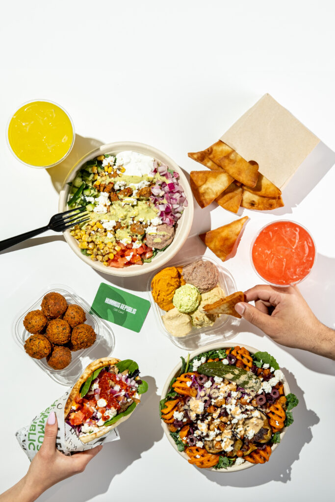 a variety of mediterranean dishes including a salad bowl, falafel, pita chips with dips, a wrap, two drinks, and a hand reaching for food on a white surface.