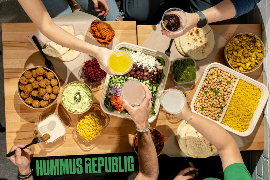 The Mental Shift That Separates People Who Think About Owning a Business From Ones Who Actually Do It overhead view of people sharing mediterranean food including hummus, falafel, chickpeas, vegetables, and drinks on a table with “hummus republic” text in the corner.