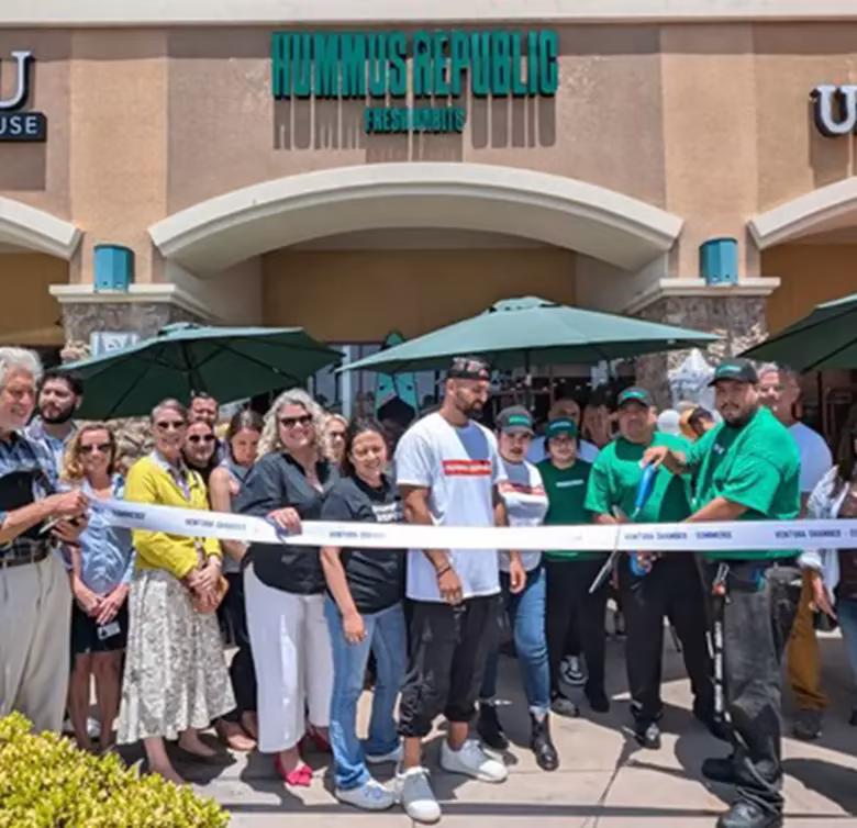 a group of people stands outside hummus republic restaurant for a ribbon cutting ceremony, with several holding the ribbon and others gathered around. a group of people stands outside hummus republic restaurant for a ribbon cutting ceremony, with several holding the ribbon and others gathered around.