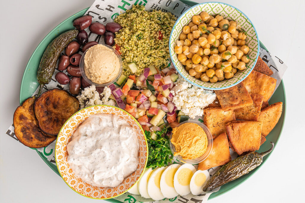a plate with pita chips, sliced egg, grilled jalapeños, olives, roasted sweet potato, tabbouleh, chickpea salad, tzatziki, hummus, feta, and diced vegetables.