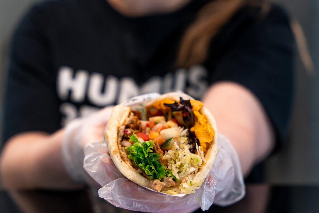 How to Build and Sell a Franchise for a Profit — What Buyers Actually Look For a person wearing gloves holds a pita wrap filled with vegetables, hummus, and other ingredients; humus is partially visible on their shirt.