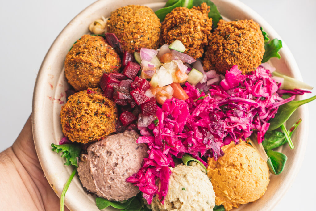 a bowl with falafel, diced vegetables, shredded red cabbage, and scoops of hummus, baba ghanoush, and another spread on a bed of mixed greens.