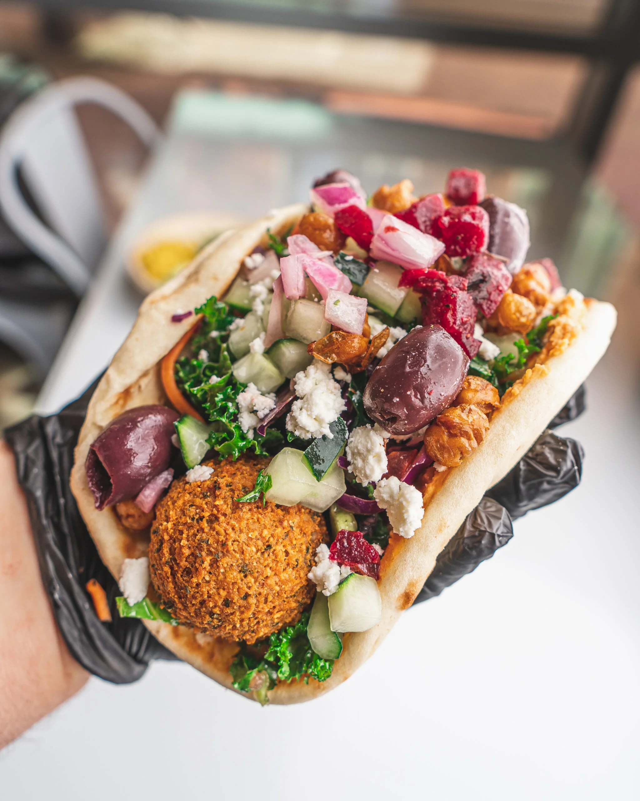 Home a gloved hand holds a pita filled with falafel, kale, cucumber, feta cheese, kalamata olives, red onion, and diced beets.