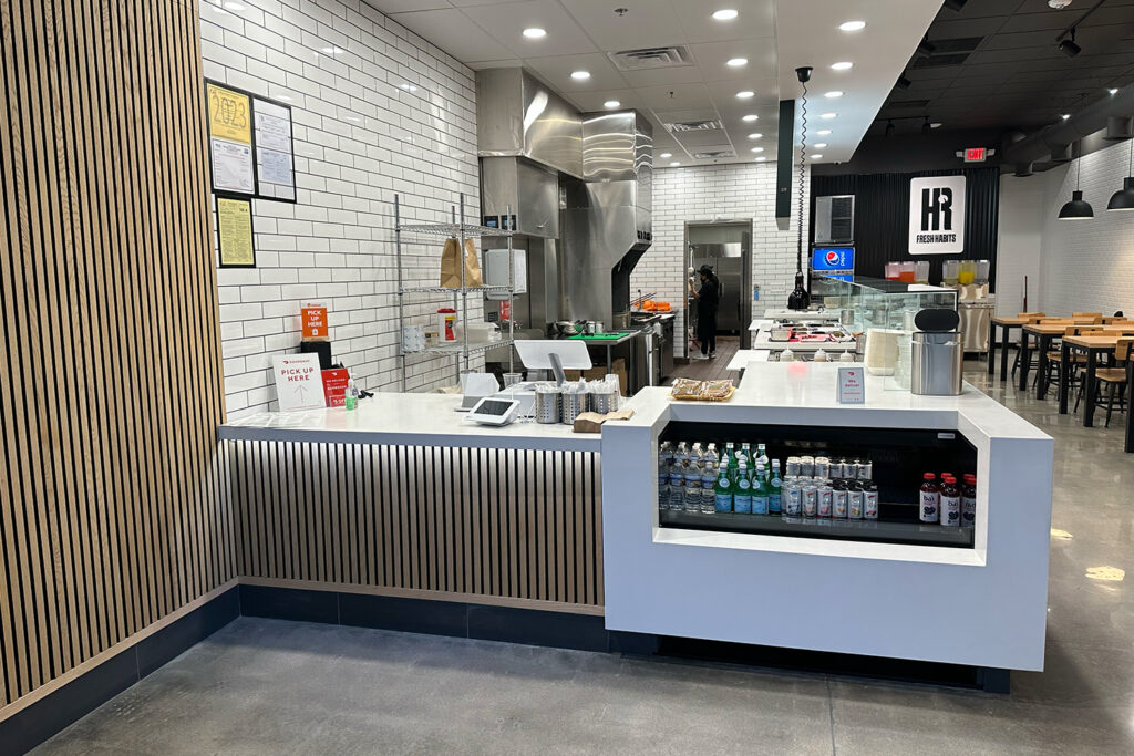 What Happens to a Neighborhood When One of Their Own Opens a Business on the Main Street modern restaurant counter with bottled drinks, a register, and food prep area in the background. dining tables and chairs are visible to the right.
