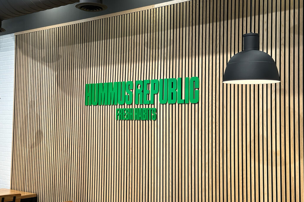 wooden slat wall with green text reading “hummus republic fresh habits” beneath a black hanging lamp.