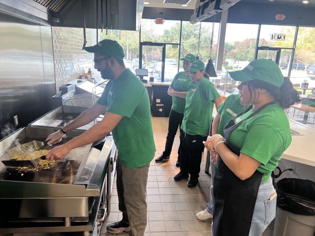What Good Unit Economics Actually Look Like in a Fast Casual Franchise four restaurant workers in green shirts and hats watch as a coworker cooks food on a grill in a modern, brightly lit kitchen.