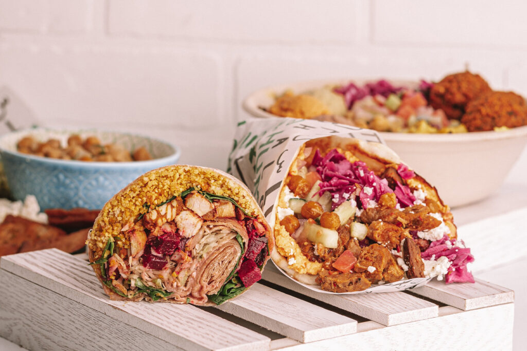 two large wraps filled with vegetables, meat, and grains are displayed on a wooden surface, with bowls of salad and falafel in the background.