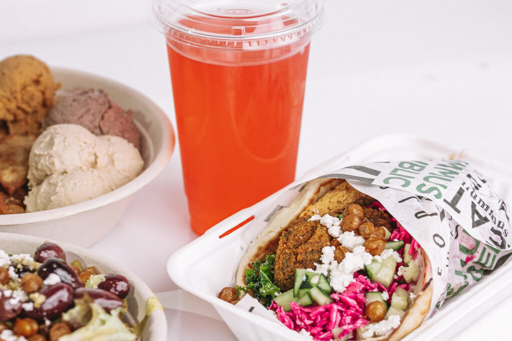 What Happens to a Neighborhood When One of Their Own Opens a Business on the Main Street a falafel wrap with vegetables and feta in a paper tray, a cup of red drink, and a bowl of assorted scoops of ice cream on a white surface.