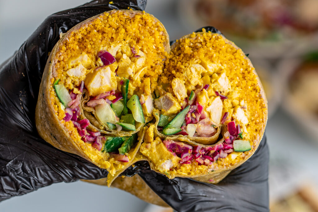 a gloved hand holds a wrap cut in half, showing yellow rice, diced chicken, red onions, cucumbers, and lettuce inside.