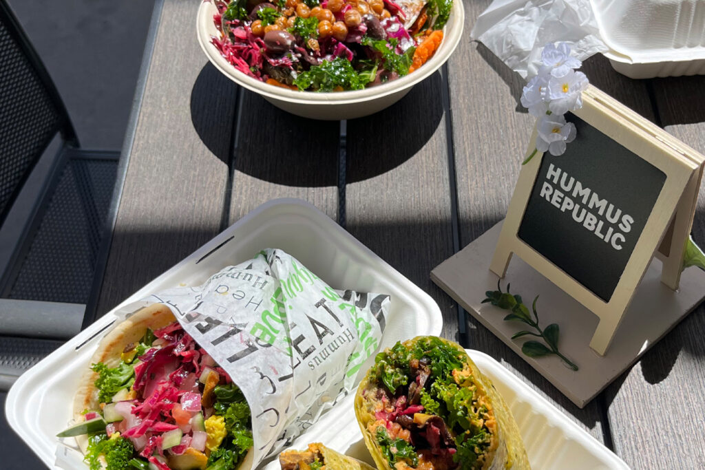 Why Food Courts and High-Traffic Corridors Are Quietly Perfect for Mediterranean Fast Casual three takeout containers on a table with a pita wrap, a salad bowl, and a wrap cut in half, next to a small sign reading “hummus republic.”.