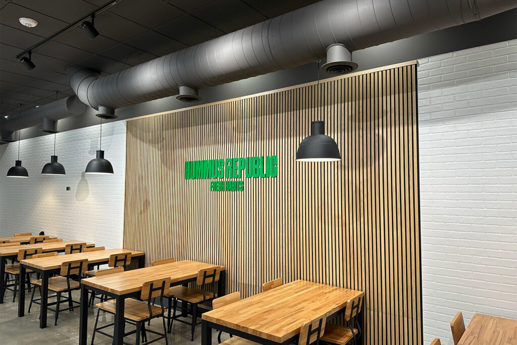 a modern restaurant interior with wooden tables and chairs, pendant lights, and a wall with vertical wood slats featuring a green unmisstekable restaurant sign.