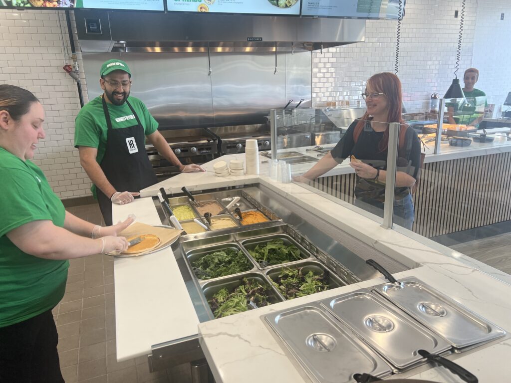 How to Build and Sell a Franchise for a Profit — What Buyers Actually Look For two employees prepare food behind a counter while a customer waits on the other side in a modern restaurant with salad ingredients on display.