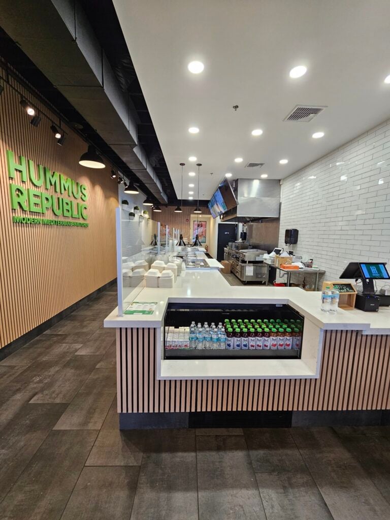 How a Focused Menu Cuts Waste, Lowers Cost, and Keeps Your Kitchen Sane modern fast casual restaurant interior with a service counter, bottled drinks display, kitchen area in back, and hummus republic signage on the wooden wall.