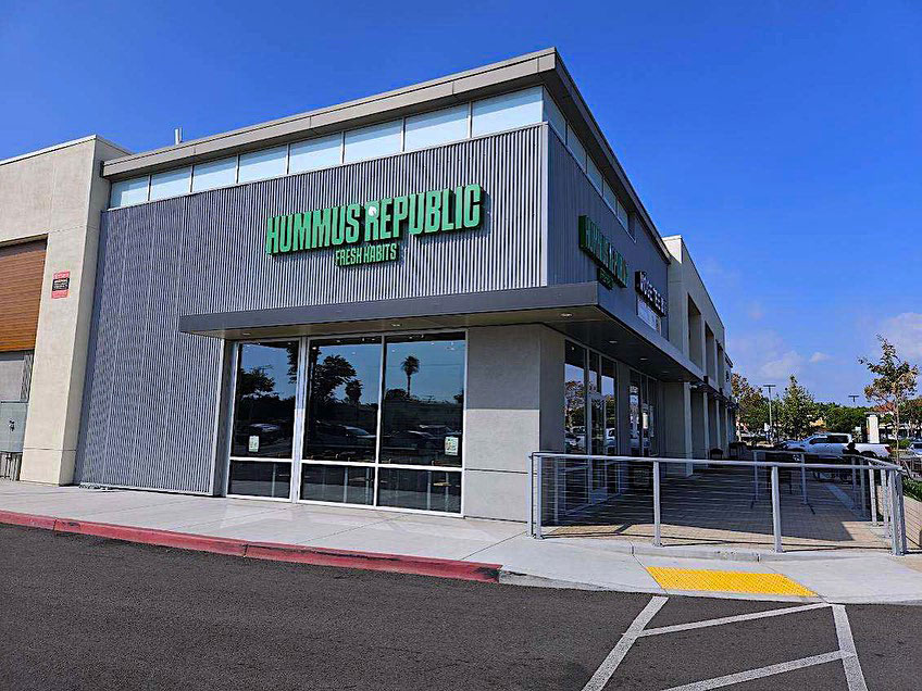 a modern corner building with large windows and a sign reading hummus republic fresh eats on the exterior, located next to a parking lot under a clear blue sky.
