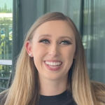 Home a woman with long blonde hair smiles at the camera, wearing a black shirt and stud earrings, with a blurred outdoor background.